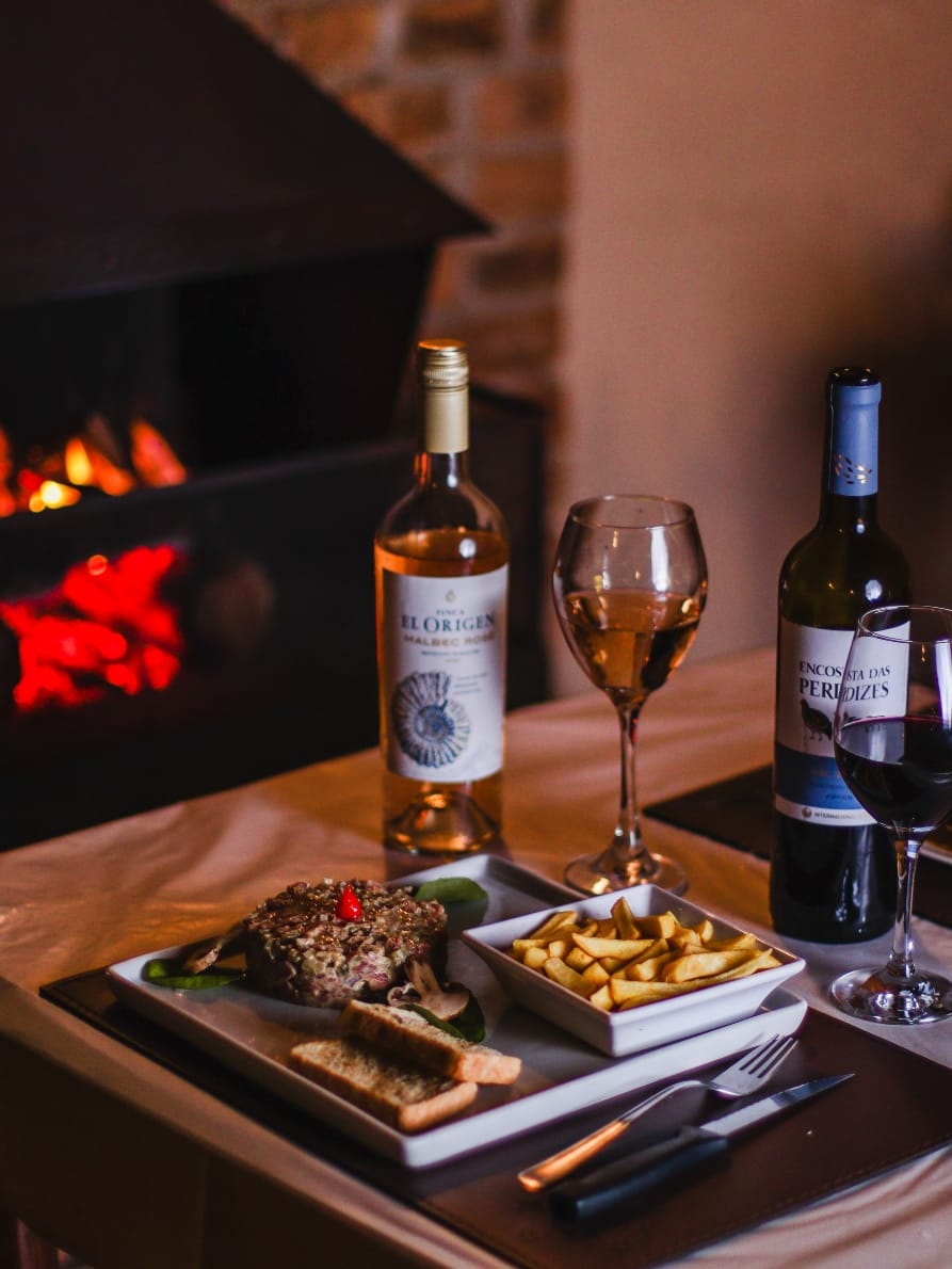 Mesa posta com steak tartare e vinhos diante da lareira